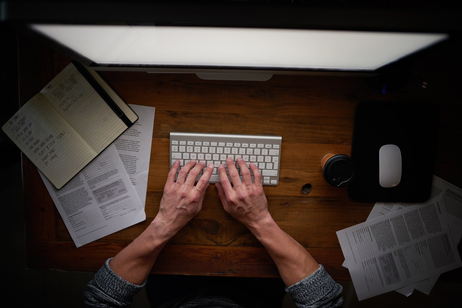 A desk at night: hands on a keyboard, an open notebook, printed pages and a lit monitor